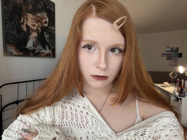 HildaGlady tem pele clara, cabelo ruivo com um prendedor de cabelo bege, veste uma blusa branca de tricô, tem olhos cinzentos e uma maquiagem discreta. Ela está olhando para a câmera com uma expressão neutra..