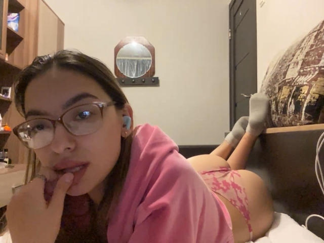 RaneeDerienzo lying on bed, pink panties, butt up, pink hoodie, glasses, finger in mouth, earbuds in, socks, mirror on wall, brown hair, looking at camera.