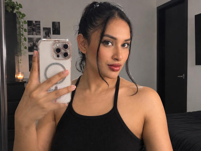 Adalina Vera mit dunklem Haar im Pferdeschwanz, trägt ein schwarzes Tanktop und hält ein Handy mit einem Ringgriff. Sie macht ein Selfie und zeigt dabei ihre Brüste und roten Lippenstift..