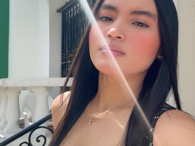 MiiaPalmer, avec de longs cheveux noirs, des joues légèrement rosées, un petit collier en or, un maquillage discret, et un reflet de soleil sur son visage, portant un haut noir à bretelles..