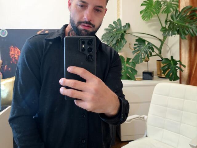 TommyDum with a black shirt, beard, and short hair taking a selfie with a black phone, showing his hand and phone in the mirror. His ass is not visible.