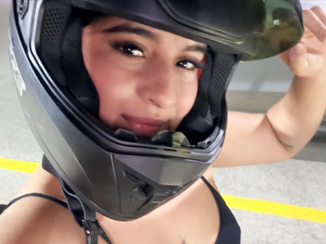 Emma Waltter, usando um capacete de motocicleta preto, segurando-o com a mão direita, mostrando seu rosto e uma blusa preta de alças finas. Seus olhos castanhos estão ligeiramente voltados para a direita..