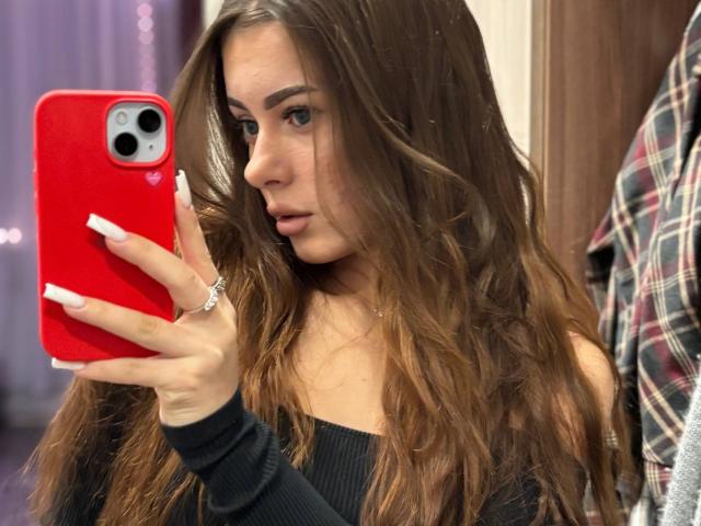LisabethLintner with long brown wavy hair, taking a mirror selfie with a red phone, showing off her white nails and a ring, wearing a black off-shoulder top.