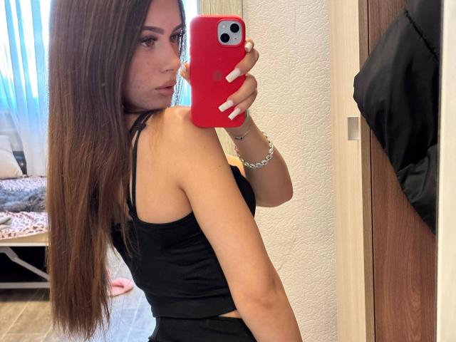Lisabeth Lintner, con cabello castaño largo, blusa negra, sosteniendo un teléfono rojo, tomando una selfie en el espejo, mostrando su trasero, mirando por encima del hombro, uñas blancas, pulsera de plata, en una habitación bien iluminada..