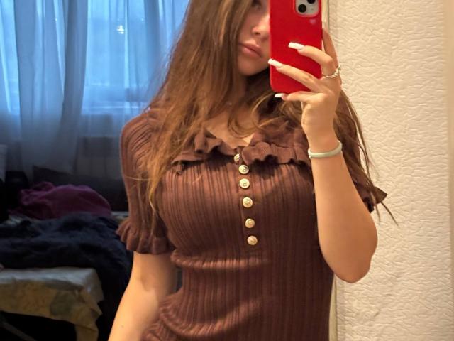 Lisabeth Lintner, con lunghi capelli castani, indossa una maglia marrone a coste con volant e bottoni, tiene in mano un telefono rosso, mostra il seno, ed è in posa per un selfie allo specchio..