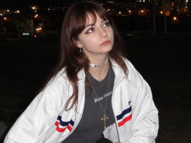 LeomaHaq with long brown hair, wearing a white jacket with red and blue stripes, grey shirt, and multiple necklaces, has a pouty expression at night.