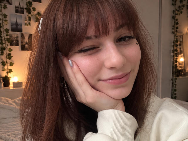 LeomaHaq has wavy brown hair, one eye closed, and is winking with a cute smile, resting her hand on her cheek. She's wearing a white sweater with long sleeves.