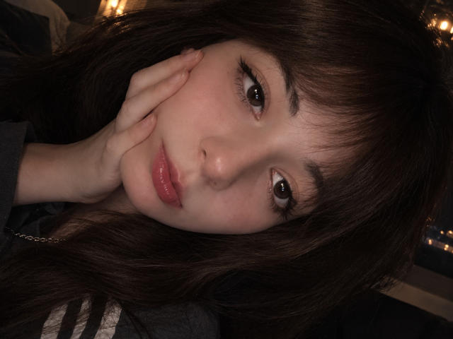 LeomaHaq has long dark brown hair, brown eyes, and glossy pink lips. She's touching her cheek with her left hand, wearing a gray shirt. Her ass is not visible.
