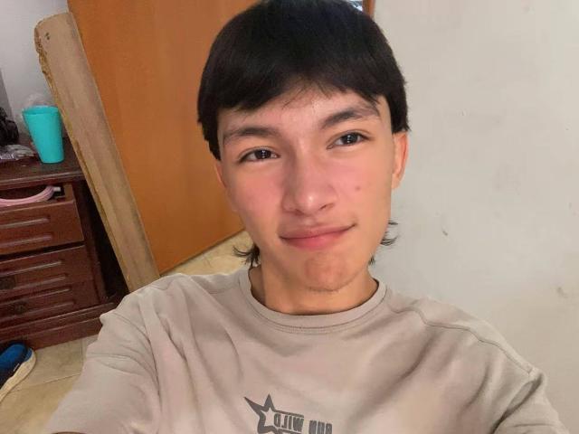 AlanMadisson, met kort zwart haar, een klein snorretje, een lichte glimlach, gekleed in een grijze shirt, die een selfie maakt. Zijn gezicht is licht rood, met een kleine puist op zijn wang..