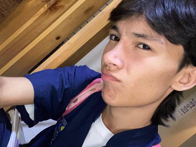 AlanMadisson with short brown hair, wearing a blue shirt and white tee, puckering his lips in a silly expression, brown eyes, tan skin, and a pink backpack strap.