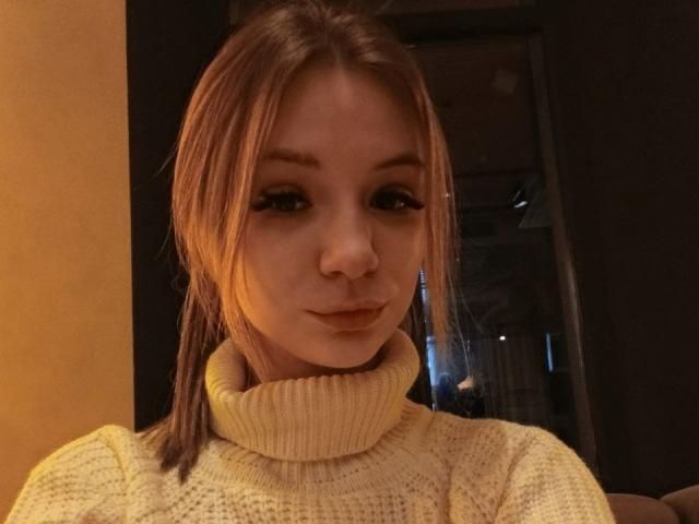 Amanda Grasy, mit braunen Haaren im Pferdeschwanz, trägt einen gelben Rollkragenpullover, hat ein hübsches Gesicht mit dezentem Make-up und eine kleine, leicht hochgezogene Nase..