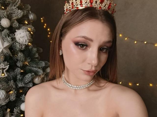 AmandaGrasy with a red and gold crown, pearl choker, and no top, showing her tits, brown eyes, and subtle makeup, in front of a decorated Christmas tree.