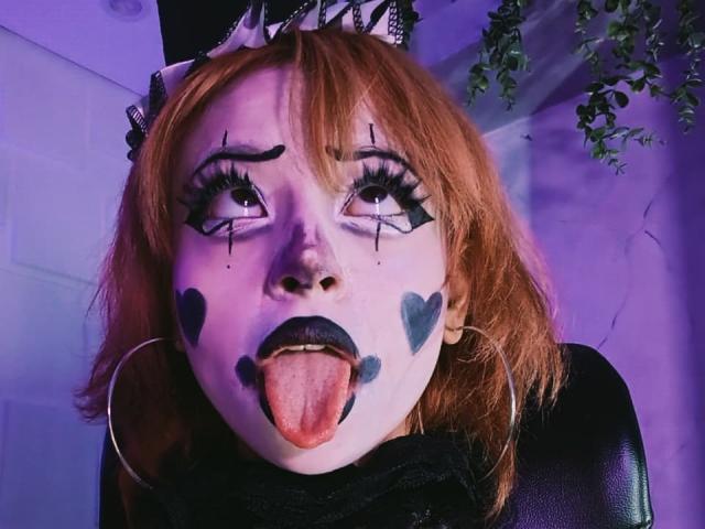 ViolettClay heeft bleke huid, oranje haar, zwarte eyeliner, hartjes geschilderd op haar wangen, zwarte lipstick, haar tong uitgestoken, en een zwarte hoge hoed met een lint. Ze draagt grote oorringen..