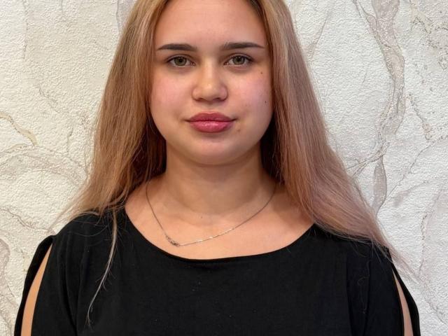Rheba Haycook mit langen blonden Haaren, einem schwarzen Oberteil, kleinen Brüsten, heller Haut, pinkem Lippenstift und einer silbernen Halskette, steht vor einer strukturierten weißen Wand..