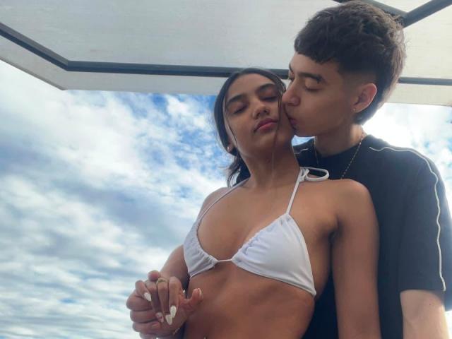 ScarlettAndMatias in a white bikini, showing small tits, with Matias kissing her neck, holding her hand, both with brown hair, under a white canopy, sky in background.