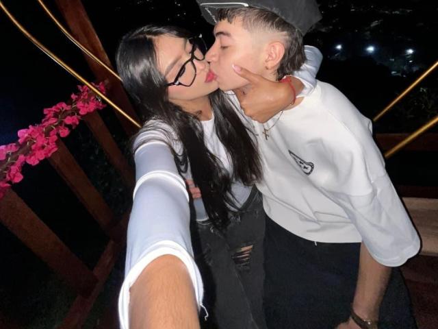 Scarlett und Matias küssen sich, ihre Hand liegt auf seiner Wange, sie trägt eine Brille, hat langes, schwarzes Haar, weiße Ärmel und schwarze Hosen. Matias trägt ein weißes Hemd, schwarze Hosen und hat kurze, blonde Haare. Im Hintergrund ist ein Holzbalkon zu sehen..