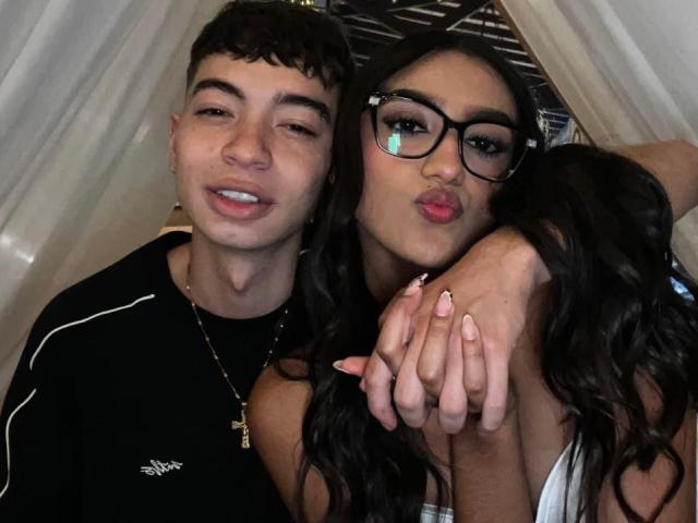 Scarlett en Matias met brillen, ondeugende lippen en lange nagels, armen om Matias heen, die lacht, beiden met donker haar, in zwart-witte kleding, binnen, in selfie-stijl..