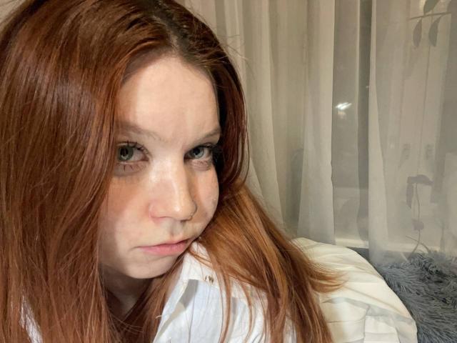 LilliamChevalier has red hair, a nose ring, and a pensive expression, with freckles on her pale skin, and is wearing a white shirt, looking slightly down.