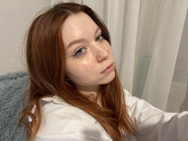 LilliamChevalier has shiny, freckled skin, long red hair, and blue eyes, wearing a white shirt, with a soft, grey fuzzy pillow behind her.