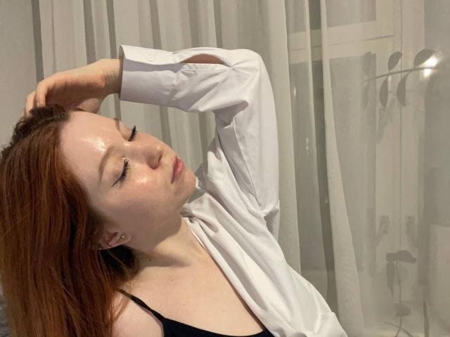 LilliamChevalier has shiny skin, red hair, and is wearing a white shirt with black straps, pouting with eyes closed, hand in hair. She's looking cute and sexy.
