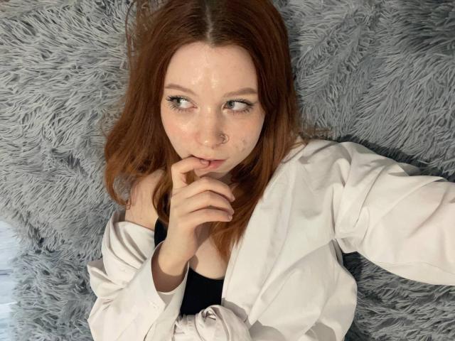 LilliamChevalier with red hair, nose ring, and white shirt, lying on a gray furry blanket, thinking with her finger near her mouth, cum on her face.
