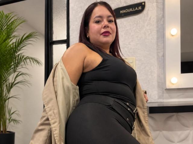 Maya Malik, con un seno di media grandezza, indossa una canotta nera e pantaloni neri aderenti, una giacca beige è tolta, guarda la fotocamera con un leggero sorriso, ha i capelli castano scuro e rossetto rosa..