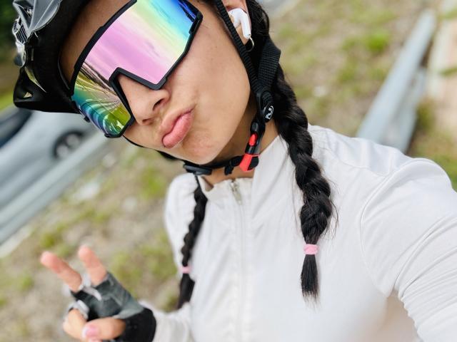 KimmyK with rainbow reflective sunglasses, black braid, white long-sleeve, black helmet, and fingerless gloves, pouting lips, wearing earbuds, outdoors, hand raised, soft pink lipstick.