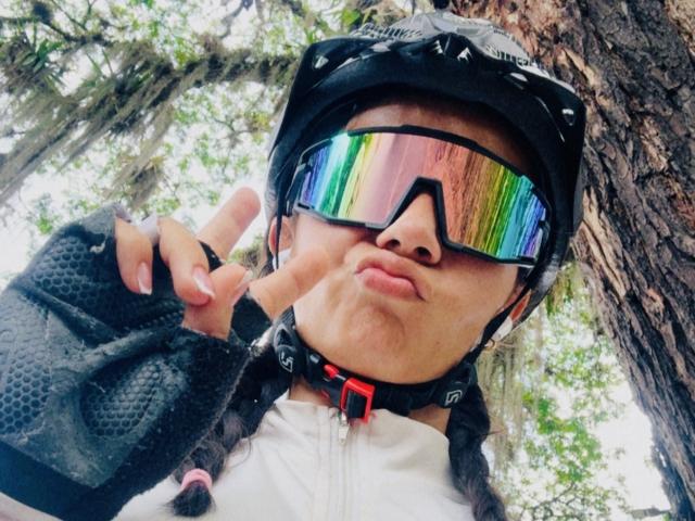 KimmyK with rainbow reflection sunglasses, black helmet, white jacket, fingerless gloves, puckering lips, making a peace sign, tree in background, braided hair, red collar, sporting a cute face.
