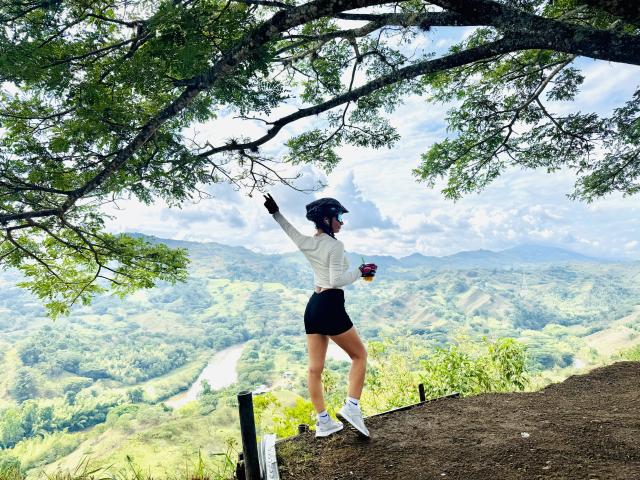 KimmyK stands on a dirt ledge, arms raised, wearing a white top, black shorts, white sneakers, and a blue cap, holding a red cup, with a mountain view.