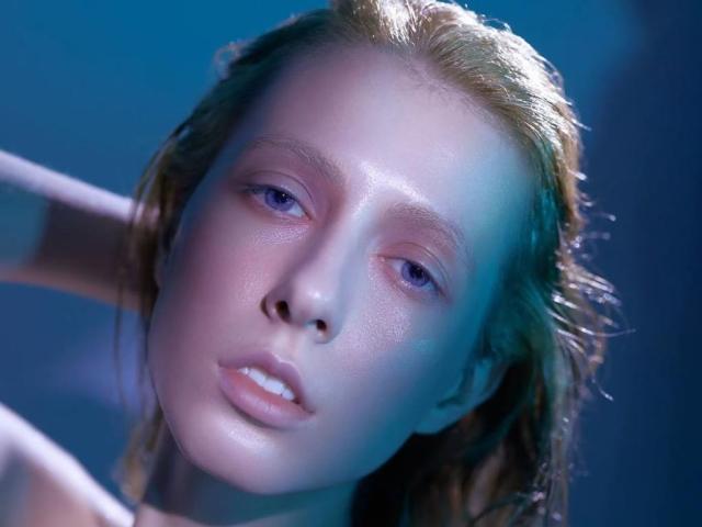 Le visage de Reanna Dreben, légèrement incliné, avec des cheveux blonds mouillés, des reflets bleus et roses sur sa peau pâle, et des lèvres fines, rosées. Ses yeux sont à moitié ouverts, et elle regarde légèrement vers le bas..