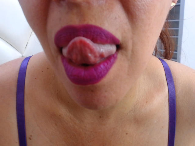 LizethBells's face with purple lipstick, tongue out, licking her upper lip. She's wearing a purple bra, visible collarbone, and a slight tan. Her tongue is wet and red.