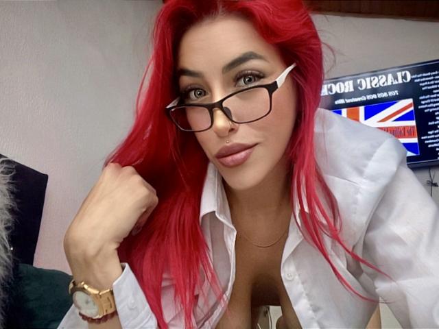 AlissaBrown with red hair, black glasses, white shirt, nose ring, and gold watch, showing her cleavage, holding hair, wearing a septum ring, and looking at the camera.
