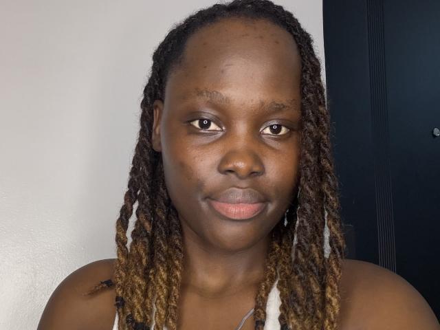 Fatma69 a la peau brun foncé, des dreadlocks de longueur moyenne avec des pointes blondes, et une expression sérieuse. Elle porte un débardeur blanc qui laisse apparaître ses épaules nues et ses fesses..