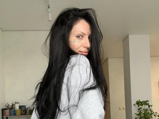 Karinisse, avec ses longs cheveux noirs, son t-shirt gris et ses petits tatouages en forme d'étoile sur la joue, regarde par-dessus son épaule, montrant son nez et un léger froncement de sourcils, dans une pièce minimaliste..
