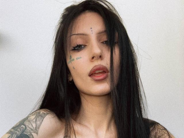 Karinisse, con i suoi lunghi capelli neri, tatuaggi sulle braccia e piccoli tatuaggi a forma di stella sulle guance, ha labbra piene, eyeliner scuro e una bocca leggermente aperta. I suoi occhi sono socchiusi..