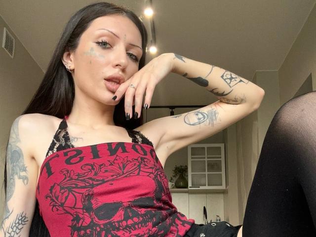 Una ragazza con tatuaggi, capelli scuri e una maglia di pizzo nero, che fa la smorfia toccandosi il mento, con una stampa di teschio rossa e nera, smalto per unghie nero e un trucco leggero, in un ambiente interno informale..