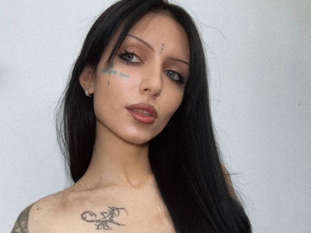 Karinisse ha lunghi capelli neri, occhi verdi, rossetto rosso, tatuaggi sul viso e sul petto e orecchini. Sta guardando la fotocamera con un leggero sorriso..