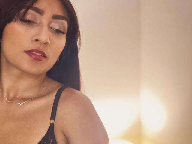 AmeliRoux with dark brown hair, red lipstick, and subtle eye makeup, wearing a black lace bra, has a silver necklace, and a soft, slightly blurry background.