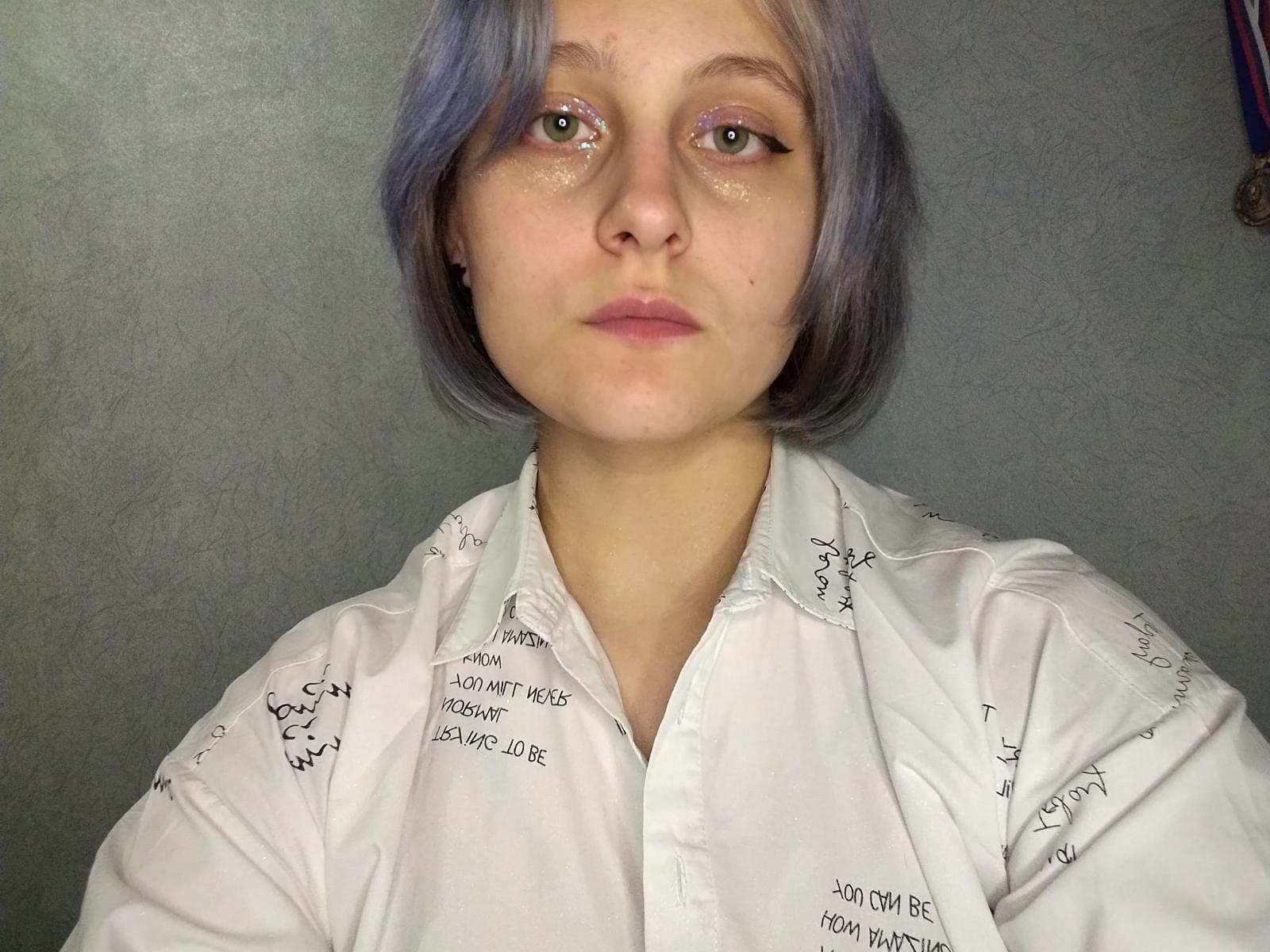 JuleDevonshire with purple-dyed hair, glittery eyes, and pink lips, wearing a white shirt with black scribble patterns, has a neutral, slightly bored expression.