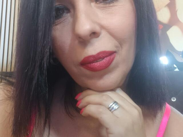 LauraSin with red lipstick, black hair, pink top, red nail polish, and silver ring, resting chin on hand, smiling slightly. Boobs partially visible. Soft, natural light.