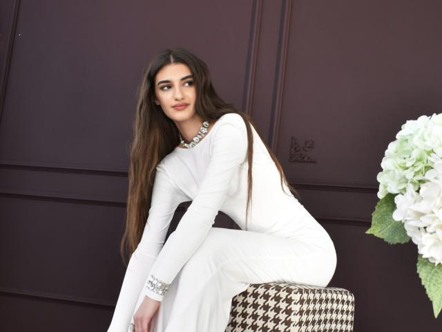 SabrinaWaynee with long brown hair, white dress, sitting on houndstooth stool, white flowers to the right, wearing silver bracelet, dark purple wall in background.
