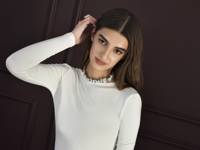 SabrinaWaynee with medium tits, wearing a white long-sleeve top and a pearl necklace, has brown hair, dark eyes, and a slight smile, touching her hair.
