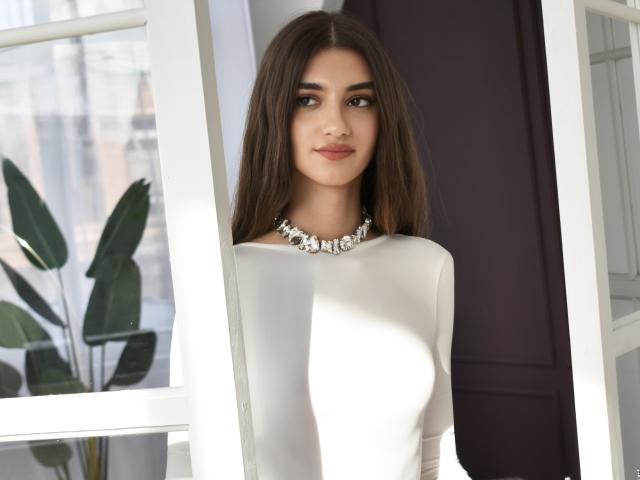SabrinaWaynee has long brown hair, a white dress, and a silver necklace, standing by a window with sunlight. Her tits are small, and she's smiling subtly.