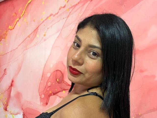 Valeria Rodriguez ha i capelli neri e lisci, rossetto rosso e indossa una maglia nera in pizzo, guardando oltre la spalla con un leggero sorriso. Ha un seno di medie dimensioni e occhi marroni..