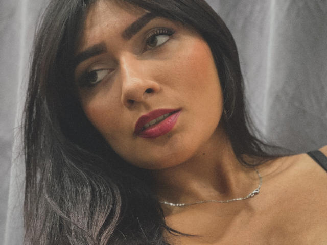AmeliRoux has tan skin, dark straight hair, and pink lipstick. She's looking to the side with a slight pout, wearing a black top and a delicate necklace.