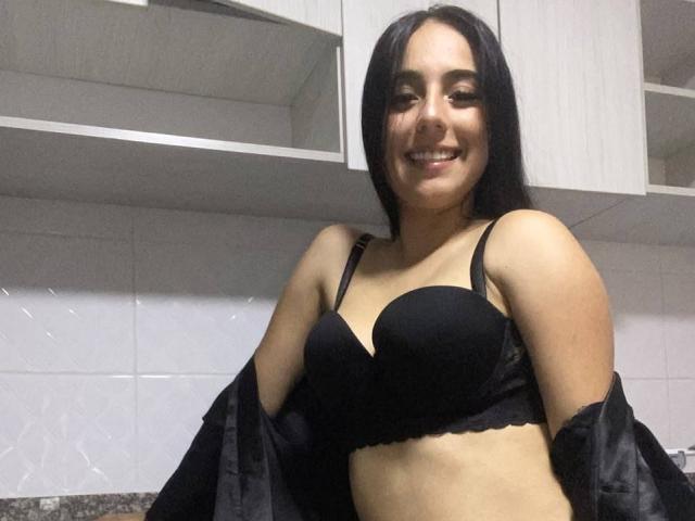 StefanyDelly with black bra, smiling, black hair, medium tits, light skin, black shirt off shoulders, kitchen background, granite counter.