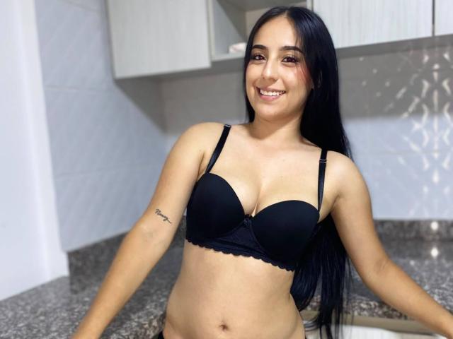 StefanyDelly with long black hair, smiling, wearing a black bra, showing her small tits and a tattoo on her right arm, standing in a kitchen with gray granite.