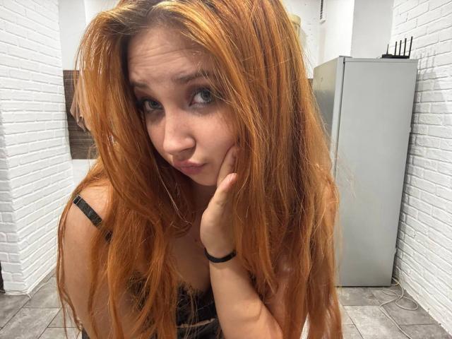 PeiSakasegawa has long, dirty-red hair, blue eyes, and is pouting with her hand on her cheek, wearing a black strap top, in a white-brick room.