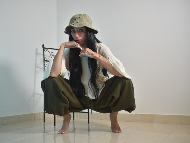 CleoZapata è accovacciata a piedi nudi, indossa un cappello a secchiello verde, una camicia bianca e pantaloni color oliva, ha lunghi capelli neri, tatuaggi sulle mani e un'espressione seria, in una stanza minimalista..