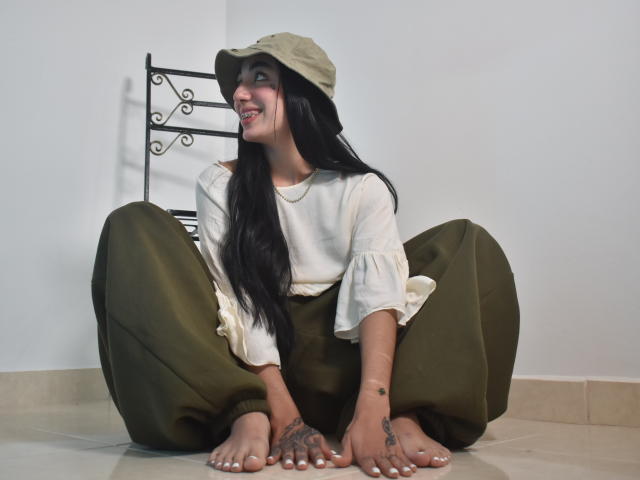 CleoZapata, assise par terre, pantalon vert, chemise blanche, chapeau seau beige, longs cheveux noirs, souriante, tatouages sur les mains, pieds nus, chaise en métal en arrière-plan, détendue, heureuse..